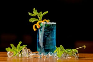 Vibrant blue cocktail garnished with mint and orange twist, set against a dark background.